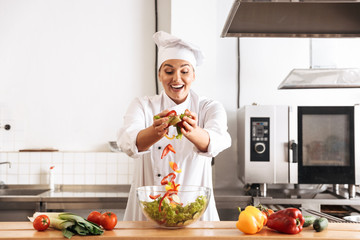 Photo of beautiful woman chef wearing white uniform making salad with fresh vegetables, in kitchen at the restaurant