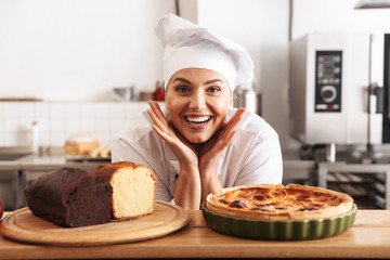 Image of adult woman chef wearing white uniform, posing in kitchen at the cafe with baked goods