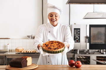 Image of delighted woman chef wearing white uniform, posing in kitchen at the cafe with baked goods