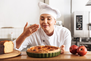 Image of pleased woman chef wearing white uniform, posing in kitchen at the cafe with baked goods