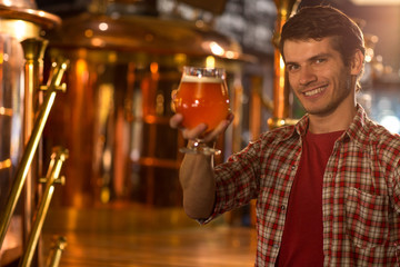 Cheerful man holding in hand and showing beer glass, while standing in beer factory. Client of brewery wearing in checked shirt looking at camera, smiling and posing.