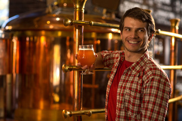 Handsome man standing with beer in hand and looking away. Man wearing in checked shirt smiling and holding glass of delicious lager. Customer of brewery posing near bronzed kettles.