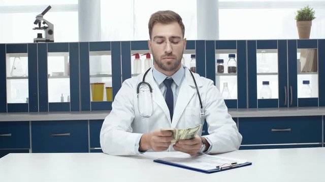 male doctor in white coat sitting at table with diagnosis, counting and putting money in pocket