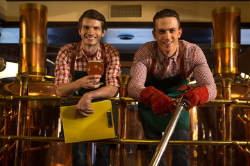 Workers of beer factory posing near bronzed equipment, kettles and pipes. Handsome brewers wearing...