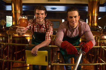 Two handsome brewers smiling, looking at camera. Men wearing in checked shirts and dark green aprons posing. Man in red gloves holding steel pipe, another worker holding folder and showing beer glass.