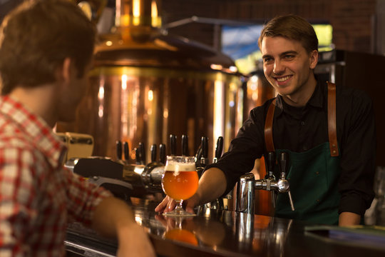 Barmen smiling, giving glass of beer to client. Man sitting at bar counter and waiting for beer. Barmen of beer house wearing in black shirt and apron looking at client.