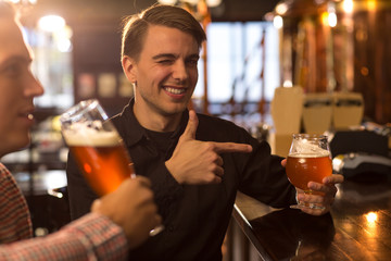 Friends drinking beer and having fun in bar. Cheerful man in black shirt winking at camera and...
