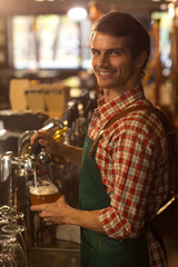 Handsome bartender looking at camera and pouring fresh, delicious beer in glass. Professional barman smiling and posing. Man wearing in checked shirt and dark green apron.