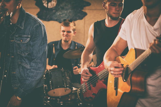 Members of rock band training on rehearsal in garage. Bass guitarist and guitarist playing on red electric guitar. Emotional drummer using drumsticks, playing behind. Singer wearing in jeans jacket