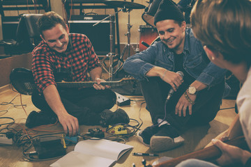 Boys of rock band sitting on floor,  and composing songs. Stylish musician wearing in black cap and jeans jacket smiling. Professional instruments, microphone and electric guitar.
