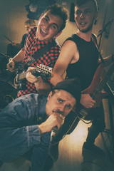 Members of youth rock band posing, looking at camera and smiling. Singer in black cap, jeans jacket holding microphone. Two cheerful  guitarists playing on electric guitar.