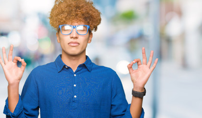 Young handsome man with afro hair wearing blue glasses relax and smiling with eyes closed doing meditation gesture with fingers. Yoga concept.