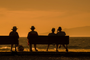 Fototapeta na wymiar People sitting on bench looking sunset on the ocean. Old men and women relaxing on a wooden sit overlooking the sea Group of man and woman enjoying the view Back side of mixed person relaxing outdoors