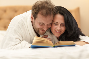 Cute wife and husband in white robes lying on bed at home, hugging and reading interesting book. Married couple spending together and enjoying relaxation. Concept of entertaining and resting.