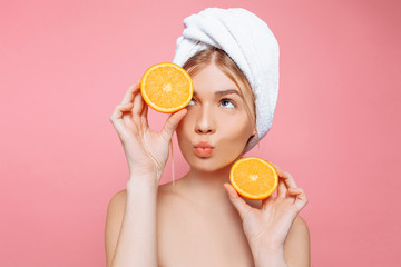 Portrait of an attractive cheerful woman with a towel wrapped around her head, holding orange slices over pink background