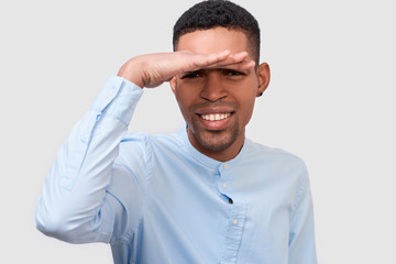 Closeup portrait of smiling African American man posing for advertisement wearing blue shirt, looking away with hand on forehead, isolated on white wall. Portrait of student male. People and emotion