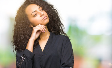 Young beautiful girl with curly hair wearing elegant dress with hand on chin thinking about question, pensive expression. Smiling with thoughtful face. Doubt concept.