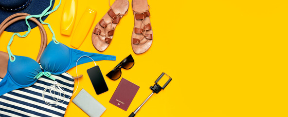 Female summer clothing accessories on yellow background. Concept of travel vacation sea. Hat swimming suit denim shorts leather sandals sunglasses sunscreen sea bag passport phone. Flat lay top view