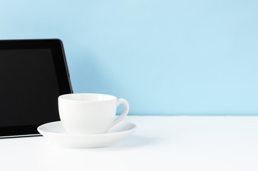 White work table with black tablet and white tea mug on a blue background. Copy space, minimal style