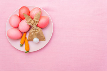 Obraz na płótnie Canvas Easter eggs concept, table arrangement decoration. Pink (rosy) eggs with bunny (rabbit) paper gift egg wrapping on plate with knitted carrots. Cloth (linen) background, top view.