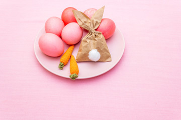 Fototapeta na wymiar Easter eggs concept, table arrangement decoration. Pink (rosy) eggs with bunny (rabbit) paper gift egg wrapping on plate with knitted carrots. Cloth (linen) background