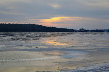 The rays of the sunset sun breaking through the gray clouds and reflected in the surface of the lake with the remains of ice and dark banks covered with forest