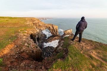 Obraz premium Man hiking and looking at The Green Bridge of Wales, Pembrokeshire Coast, West Wales, UK