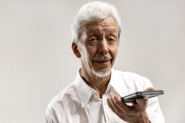 Indoor portrait of attractive senior man isolated on gray background, holding blank smartphone, using voice control, feeling happy and surprised. Human emotions, facial expression concept.
