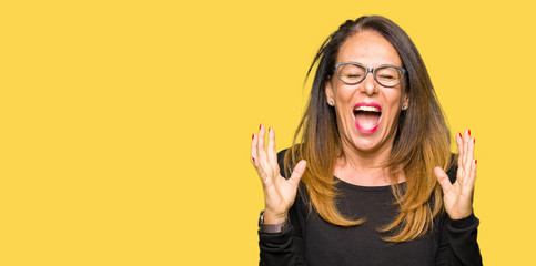 Beautiful middle age woman wearing glasses celebrating mad and crazy for success with arms raised...