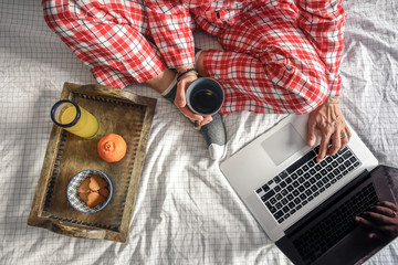Female crossed legs close-up in bed Feet of a young lady with socks and natural breakfast in bedroom Woman working at home with laptop. Orange juice and cookies on a wooden tray Young lady with a cup 