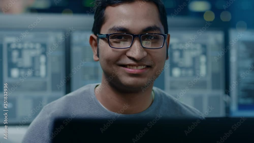 Fototapete Portrait of a Smart and Handsome Software Developer Wearing Glasses Working on a Personal Computer, Smiles at the Camera. In the Background Unfocused Personal Computers with Screens in Technical Room