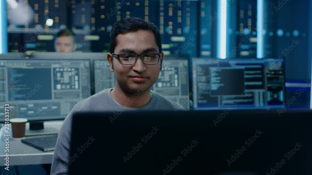 Leinwandbilder Portrait of a Smart and Handsome Software Developer Wearing Glasses Working on a Personal Computer, Smiles at the Camera. In the Background Unfocused Personal Computers with Screens in Technical Room