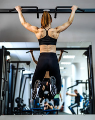 Fototapeta premium Muscular, athletic girl, bodybuilder in sportswear pulling up on a horizontal bar in front of the mirror at gym. Back muscles of a sporty girl tightening and looking at herself in the mirror.