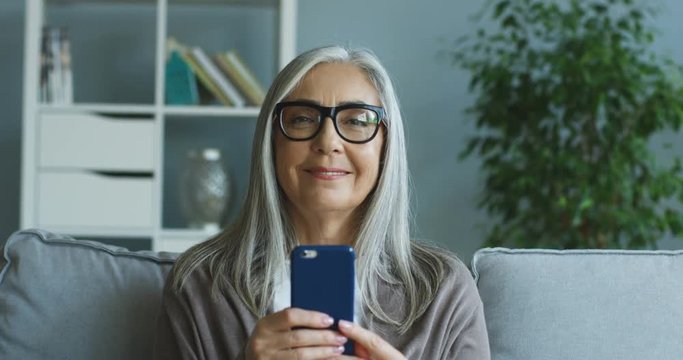Portrait of the senior Caucasian woman in glasses tapping on the smartphone and then smiling to the camera at home. Close up.