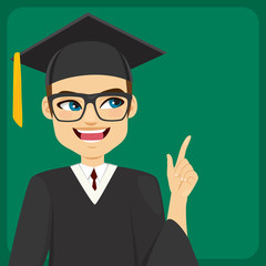 Male student with mortarboard cap and graduation gown pointing with finger to green chalkboard