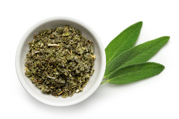 Dried rubbed sage in a white ceramic bowl next to fresh sage leaves isolated on white from above.