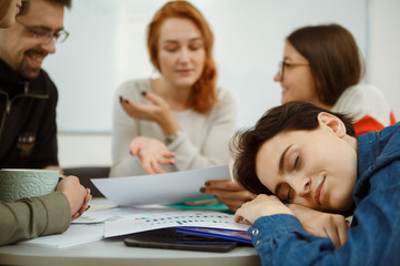 Brunette girl with short dark hair sleeping during lection in university. Female student resting after night. On background young teacher explaining something.