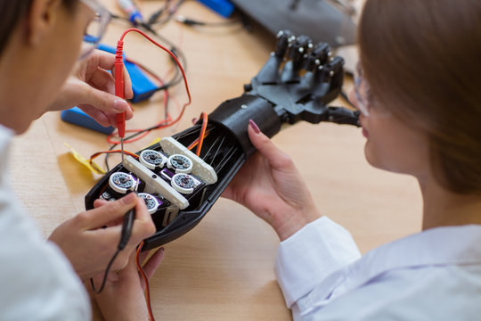 Close up of two female engineers using voltage and current tester at plastic hand. Scientist, wearing in white uniform, inspect part on which passing electricity at prototype of plastic hand of robot.