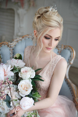 Gentle portrait of an elegant blonde bride with a bouquet of flowers in her hands in a light pink dress in the interior.