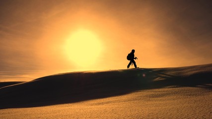 Fototapeta na wymiar photographer traveler in winter goes on snowy ridge in rays of yellow sunset. mountaineer with camera and tripod is walking in snow along top of holom.