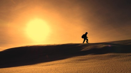 photographer traveler in winter goes on snowy ridge in rays of yellow sunset. mountaineer with camera and tripod is walking in snow along top of holom.