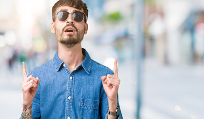 Young handsome man wearing sunglasses over isolated background amazed and surprised looking up and pointing with fingers and raised arms.