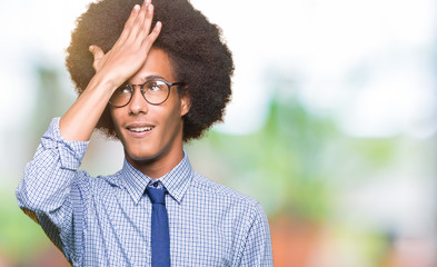 Young african american business man with afro hair wearing glasses surprised with hand on head for...