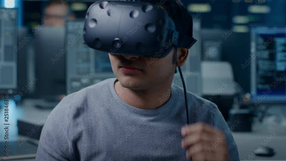 Poster Portrait of Software Delevoper Puts On Virtual Reality Headset Holds Controllers and Starting Programming in VR. In the Background Technology Developing Studio with Computers and Monitors