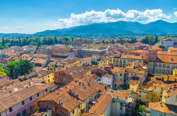Fototapeta na wymiar Aerial top panoramic view of historical centre medieval town Lucca with old buildings, typical orange terracotta tiled roofs and mountain range, hills, blue sky white clouds background, Tuscany, Italy