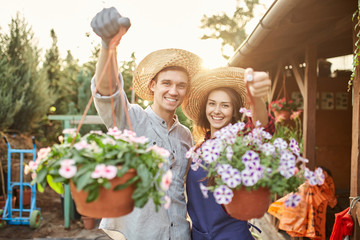 Happy guy and girl gardeners in a straw hats hold pots with wonderful petunia in the garden on a...