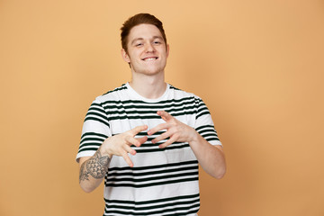 Smiling red-haired stylish guy in a striped shirt with tattoo on his hand is posing on the beige...