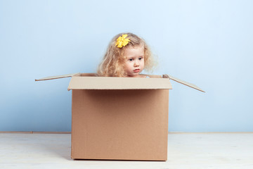 Little curly girl with yellow flower on her hair sits in the cardboard box on the background of blue wall.