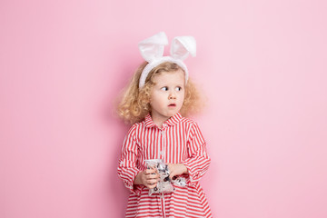 Lovely little girl in a striped red and white dress and bunny ears on her head is standing against a pink wall and eating some sweets from little packet in her hands
