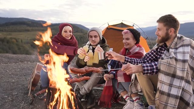Group of happy friends clining by bottle of beer while cooking sausages on the fire in the mountain.